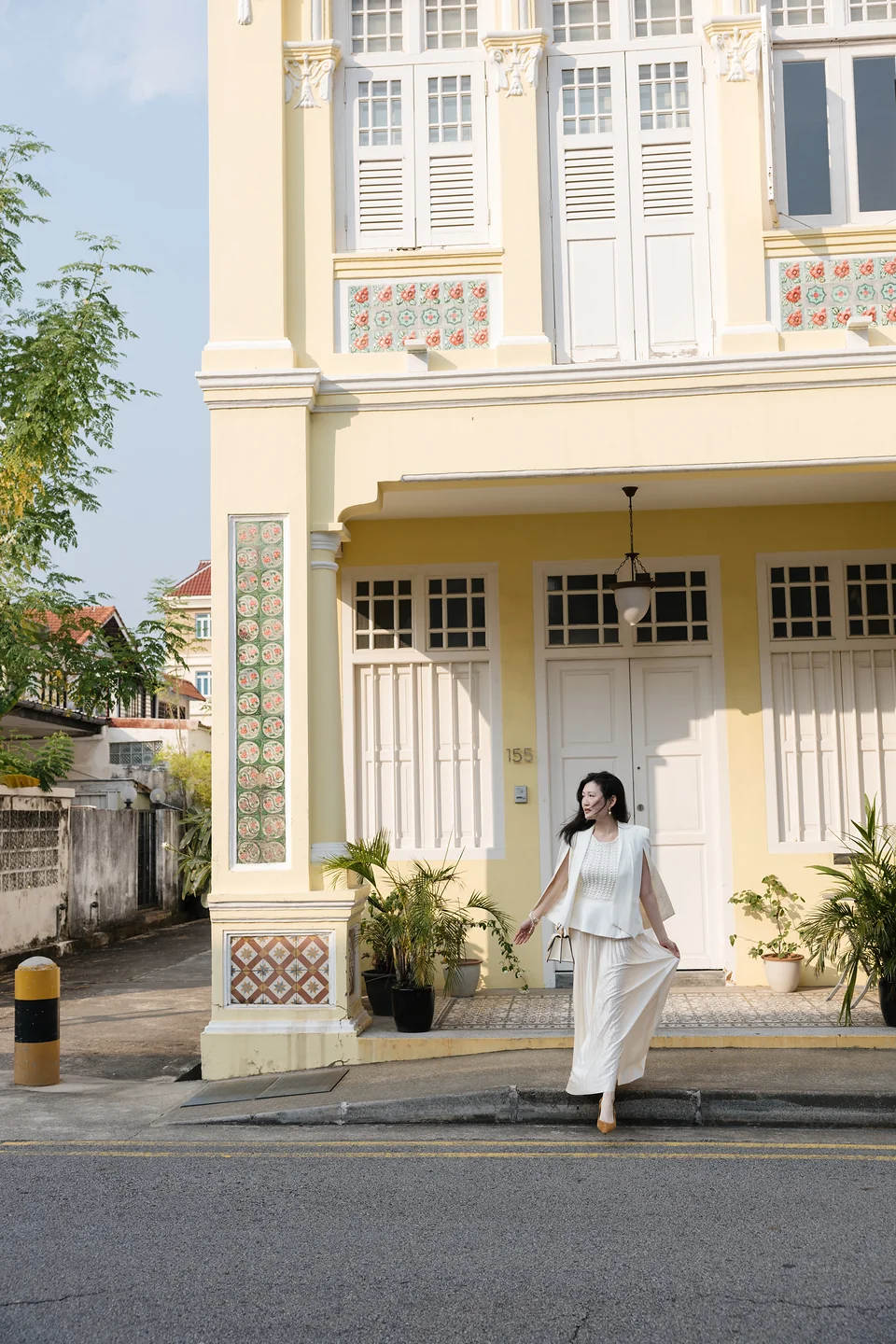 Ki beside a pale yellow heritage house with white shutters, floral tiles and decorative columns
