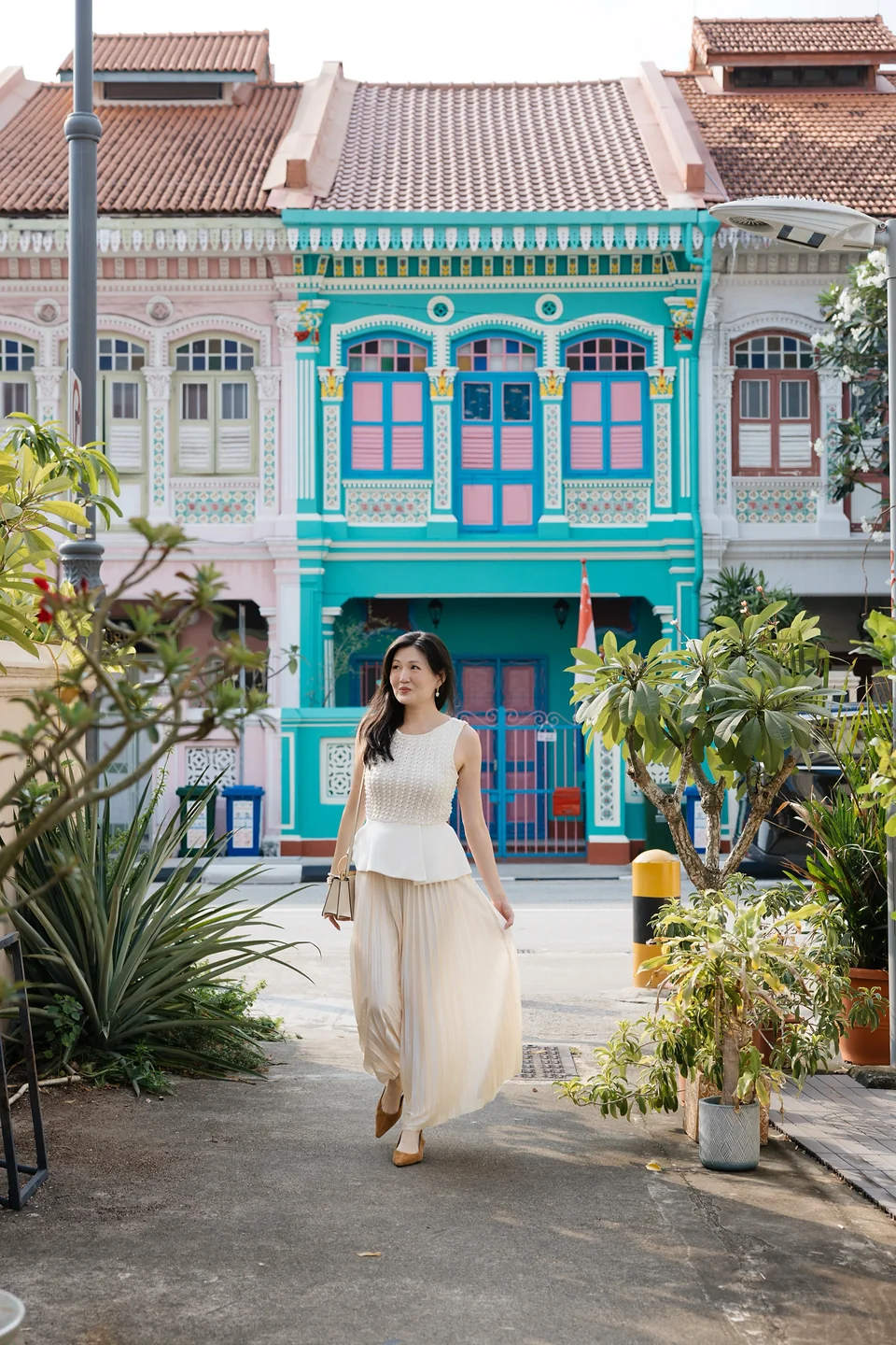 Ki walking in a plant-framed alley with a turquoise terrace house behind her