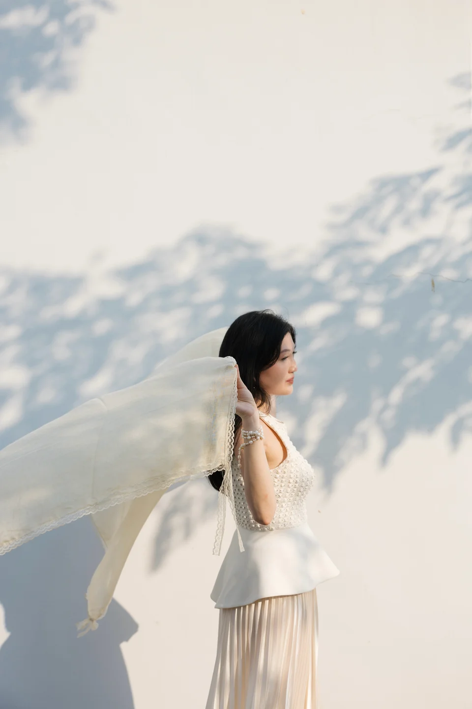 Ki holding a flowing ivory scarf against a sunlit wall with leaf shadows